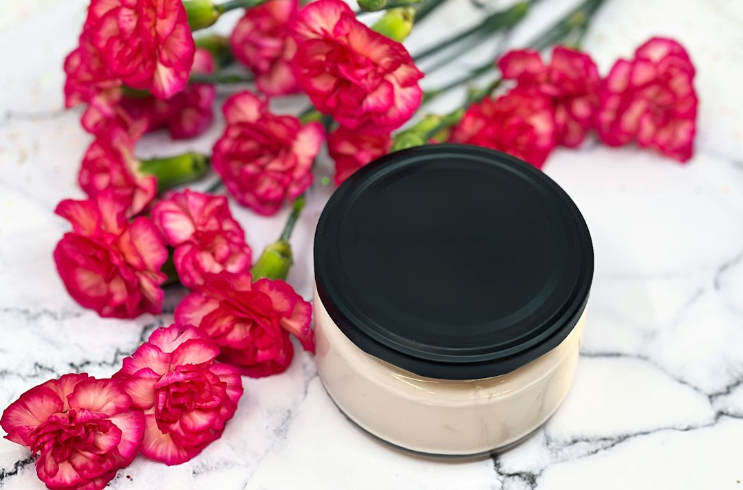 A cosmetic cream jar with a black lid arranged beside pink flowers on a marble surface.